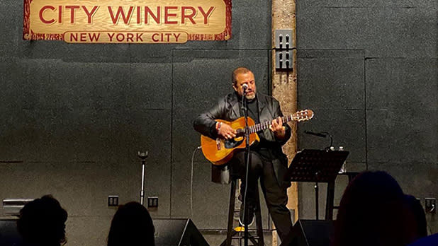 A man in a black blazer and pant set, sitting on a stool and playing the guitar at City Winery in New York City