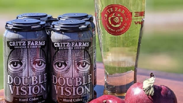 Four back of black cider cans that say "Double Vision" next to a tall glass of light cider and red apples