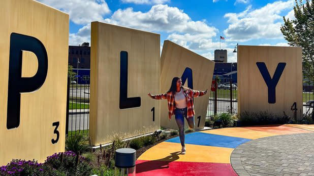 A woman jumps on a colorful sidewalk in front of giant Scrabble letters that spell out the word "play"