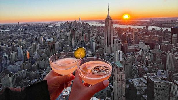 Two people "cheers" two pink cocktails in front of an aerial view on the New York City skyline at sunset