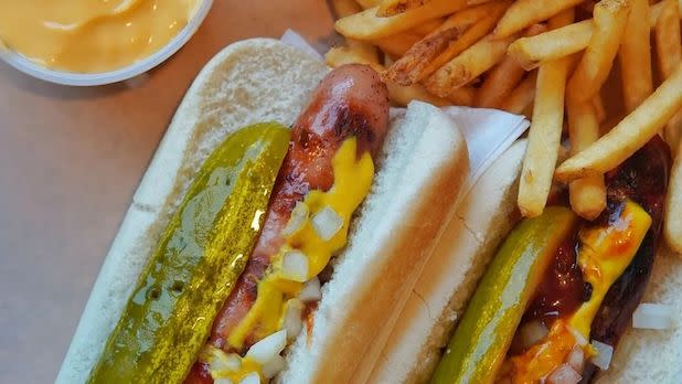 Two hot dogs with pickles, mustard, and onions on a plate with fries and a cup of cheese