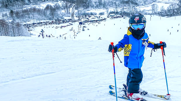 A child in a blue and yellow jacket and helmet with goggles is standing on skis and holding ski poles at the top of one of the Dry Hill Ski Area's slopes with a view of the resort and snow-covered trees below in the distance