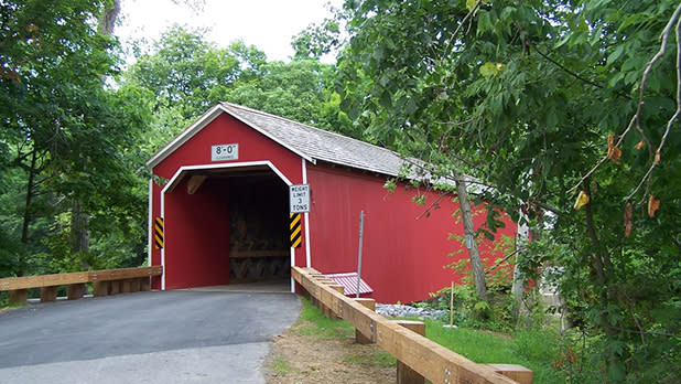 A blacktop road leads into the red and white trim portal of the Eageville Covered Bridge