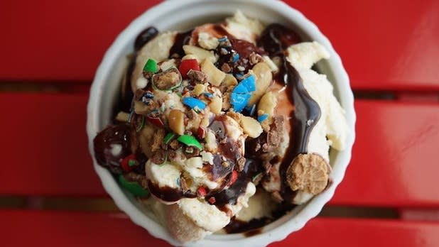 Overhead view of ice cream topped with nuts, crushed m&ms and hot fudge on a red table