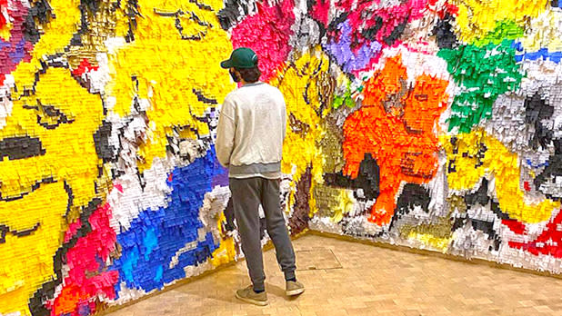 A man in gray pants and a cream sweater looking at a bright multicolored wall in El Museo Museum