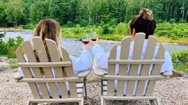 Two women sitting in beige Adirondack chairs cheers wine glasses as they look out at a creek and forest