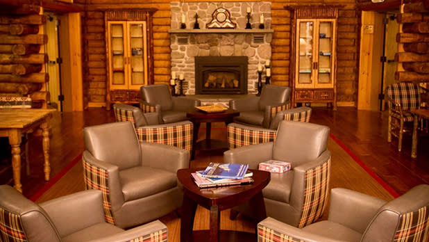 Cozy log cabin living area with six plush, plaid-accented armchairs arranged around a wooden table. A stone fireplace and warm lighting create a rustic, inviting ambiance at the Emerson Resort and Spa.