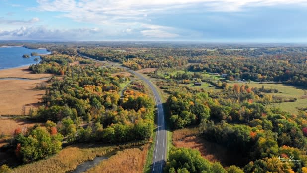 Aerial view of Route 12 winding along the St. Lawrence River through colorful autumn forest and farmland, beneath a bright, cloud-filled sky.