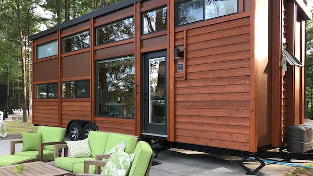 exterior of a tiny house with dark brown wood and windows at A Tiny House Resort in the Catskills. Green outdoor couches and a wood table sit in front of the house.