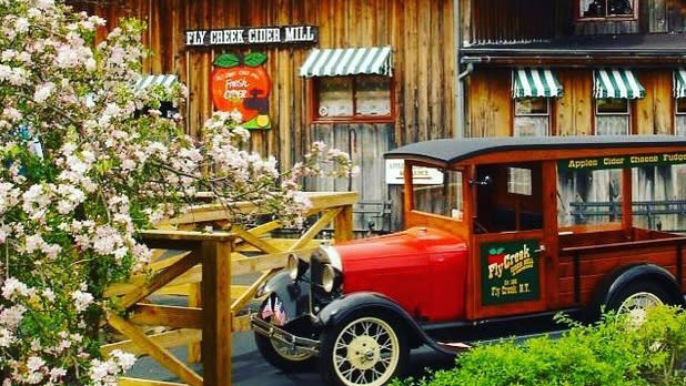 A vintage red car sits in front of the Fly Creek Cider Mill building with brown wooden paneling