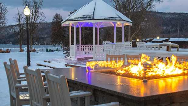 Snowy outdoor scene with a lit fire pit surrounded by chairs in the foreground. A gazebo is in the background, near a lake and a snow-covered landscape at Fort William Henry Hotel.