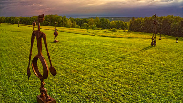 Sculptures scattered around a green field at Griffis Sculpture Park with the sun setting behind the clouds in the distance