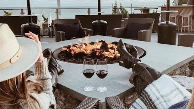 Two women in black heeled boots prop their feet up next to two glasses of red wine on the stone fire pit looking out at the ocean