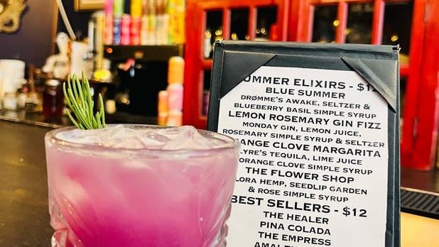 A purple elixir with a green garnish at a bar next to a "Summer Elixirs" stand-upmenu