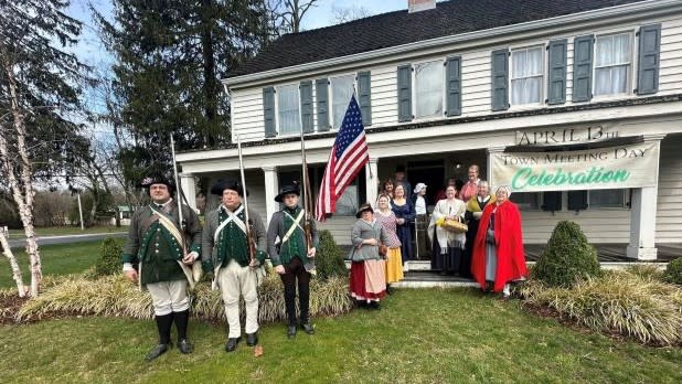 a group of reenactors stand at in front of a white house with a banner that reads "April 13" and "Town Meeting Day Celebration"
