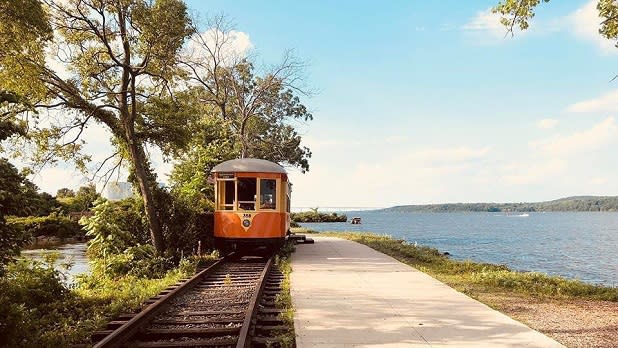 An orange and yellow trolley sits on a track next to the river, mountains, and green vegetation