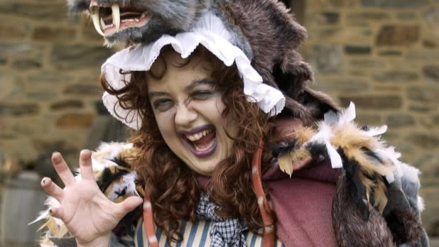 Smiling woman in a rustic wolf costume, with a wolf puppet perched on her head, playfully raises clawed hands outside Philipsburg Manor during their Halloween festivities.