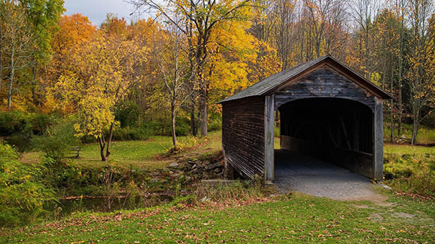 The dark wood exterior of Hyde Hall Covered Bridge with a small pond to the left and yellow, orange, and green trees all around