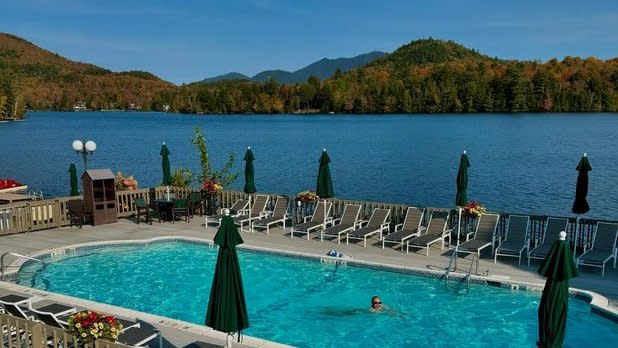 a view overlooking a blue in-ground pool surrounded by lounge chairs with Mirror Lake and the Adirondack Mountains with fall colors in the background