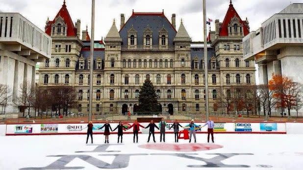 people holding hands in a line on an ice rink in front of the New York Capital building