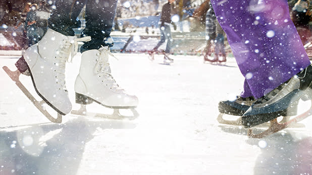 A pair of skaters skating on an ice rink. One skater has black tights and white skates and the other has purple pants and black and silver skates