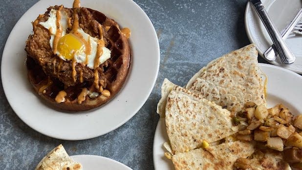 two plates of food, one with a large waffle, crispy chicken, and egg, and the other with a breakfast quesadilla and home fries.