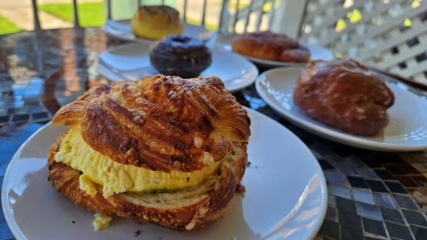 A close-up of a breakfast sandwich on a white plate, featuring a Dutch Knuckle with crustless quiche. Blurred background shows other pastries on plates outside Early Dawn Confections in Saranac Lake.