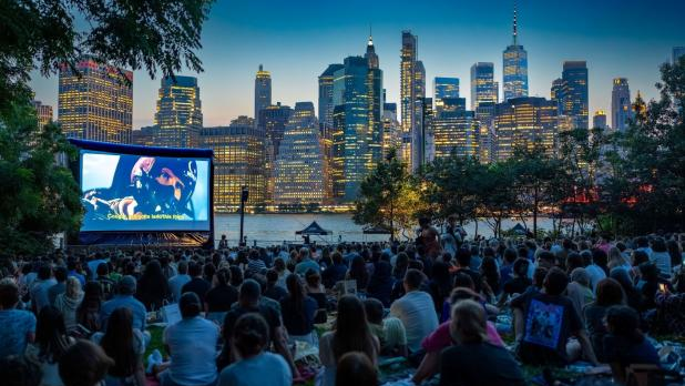 Outdoor movie night in Brooklyn Bridge Park with a crowd sitting on blankets, watching a large screen. The New York City skyline glows in the twilight.