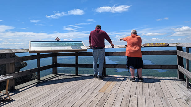 Two people stand on a wooden observation deck at Whiteface Mountain overlooking a vast, lush valley and distant mountains under a bright blue sky.