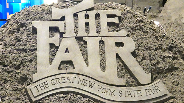 A detailed sand sculpture carved with the bold lettering "THE FAIR" above a banner reading "THE GREAT NEW YORK STATE FAIR."