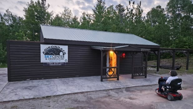 A person on a mobility scooter approaches a wooden building at dusk, labeled "Adirondack Sky Center & Observatory," surrounded by trees and a soft sky.