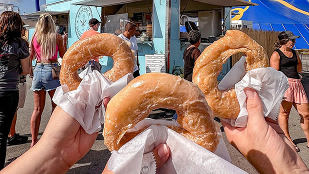 Three hands holding up glazed doughnuts from Peachey’s Baking Company at the New York State Fair.