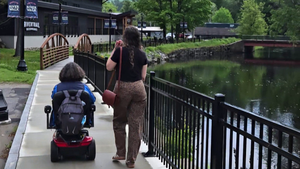 A person in a mobility scooter and a woman walking alongside on a Saranac Lake's Riverwalk path with ornate bridges, green trees, and calm water, creating a serene setting.