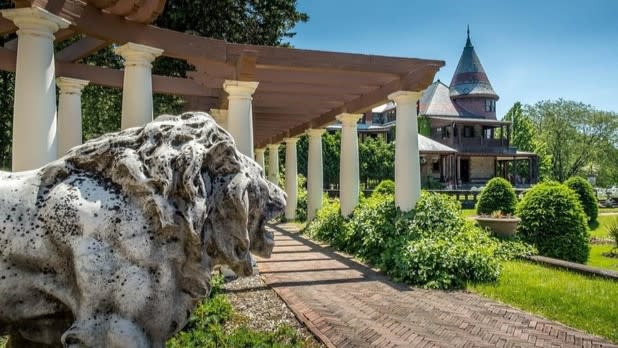 A lion statue perched in front of a brick walkway lined with pillars with the Sonnenberg Mansion in the background.
