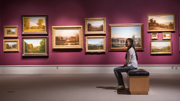 a woman sits on a bench in an art gallery with purple walls filled with Hudson River School paintings