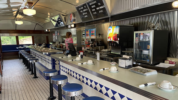 Retro diner interior with chrome stools, tiled counter, coffee cups, menu board, and a woman behind the bar, creating a cozy nostalgic mood at Chef Darrell's Mountain Diner.