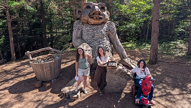 Three people stand in front of a large wooden troll sculpture in a forest at the Wild Center in Tupper Lake. One person uses a red mobility scooter. The mood is joyful and adventurous.