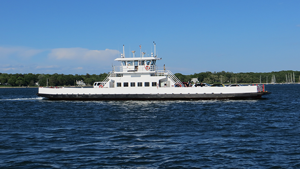North Ferry cruises across deep blue water on a sunny day, with shoreline trees, sailboats, and a clear blue sky behind it.