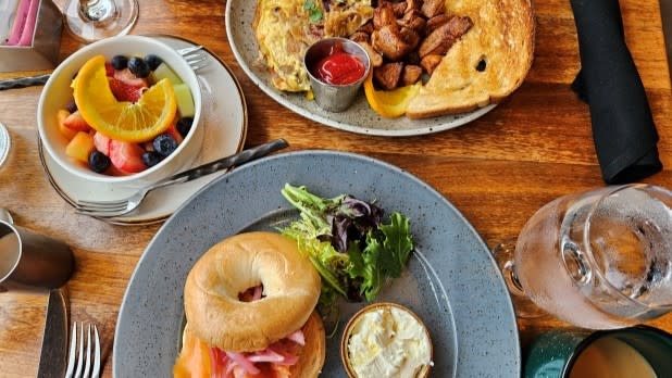 A breakfast spread on a wooden table includes a fruit bowl with orange slices, berries, an omelet with toast and potatoes, and a bagel with salad and cream cheese at the Campfire restaurant in Hotel Saranac.