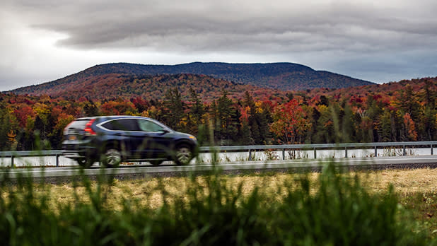 A black SUV speeds along scenic Route 28 beside Durant Lake, framed by vivid autumn forest and a broad mountain beneath dramatic gray storm clouds outside Blue Mountain Lake.