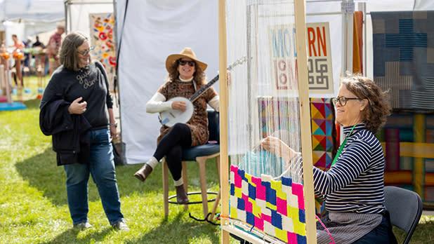 A woman weaves colorful patterns on a loom at the M&T Bank Clothesline Art & Fine Craft Festival . Another woman plays a banjo, creating a lively and creative atmosphere.
