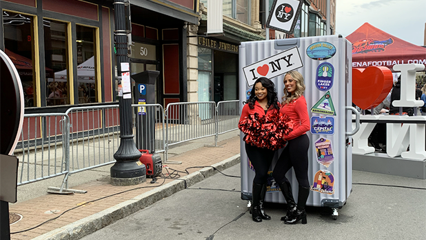 Two women pose in front of an oversized suitcase at the I LOVE NY booth