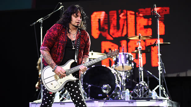A musician with long dark hair plays a white electric guitar on a stage with a drum set behind him and a backdrop displaying the band name "Quiet Riot" in red lettering at the Great New York State Fair.