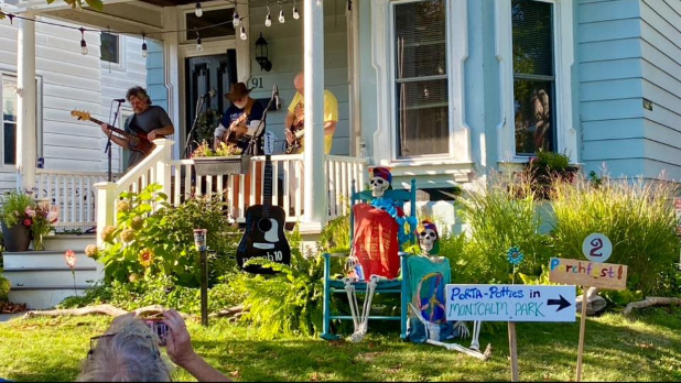 Colorful front porch with musicians performing during Oswego Porchfest, garden decorations and skeletons, bright flowers, and a sign for a “Peeps-R-Us” memorial park.