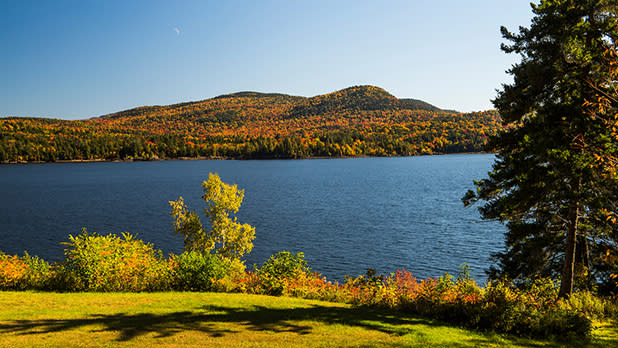Indian Lake framed by trees and a grassy shore, with colorful forested hills and clear blue sky reflecting a calm fall scene.