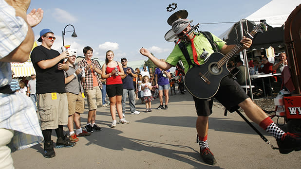 Bandaloni, the one-man band street performer, wearing a bright green shirt, red tie, and a helmet full of instruments plays a black acoustic guitar and harmonica while kicking out his leg for a cheering crowd at the Great New York State Fair.