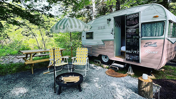 Two yellow lawn chairs surround a fire pit outside a white and pink vintage camper at Boheme Retreats in the Catskills