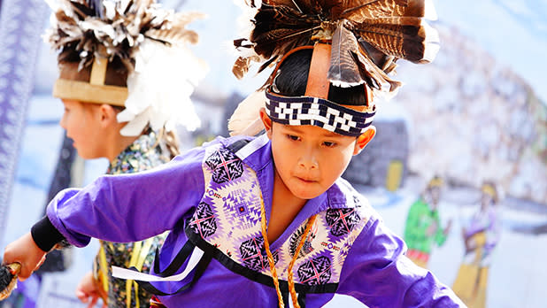A young child wearing traditional Indigenous regalia, including a feathered headdress and a purple patterned tunic, performs a dance with focused intensity at the Great New York State Fair.