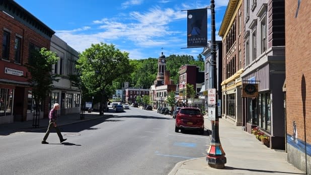 A person crosses Main Street in Saranac Lake on a sunny day. The street is surrounded by historic brick buildings, a leafy tree, and a distant clock tower.