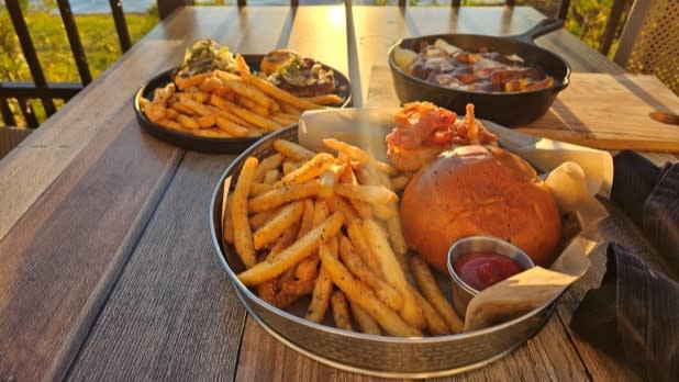 A wooden table on the outdoor patio of Saranac Lake's Lakeside Drift Restaurant with plates of burgers, fries, and a skillet dish. Sunlight casts warm shadows, creating an inviting and relaxed dining atmosphere.